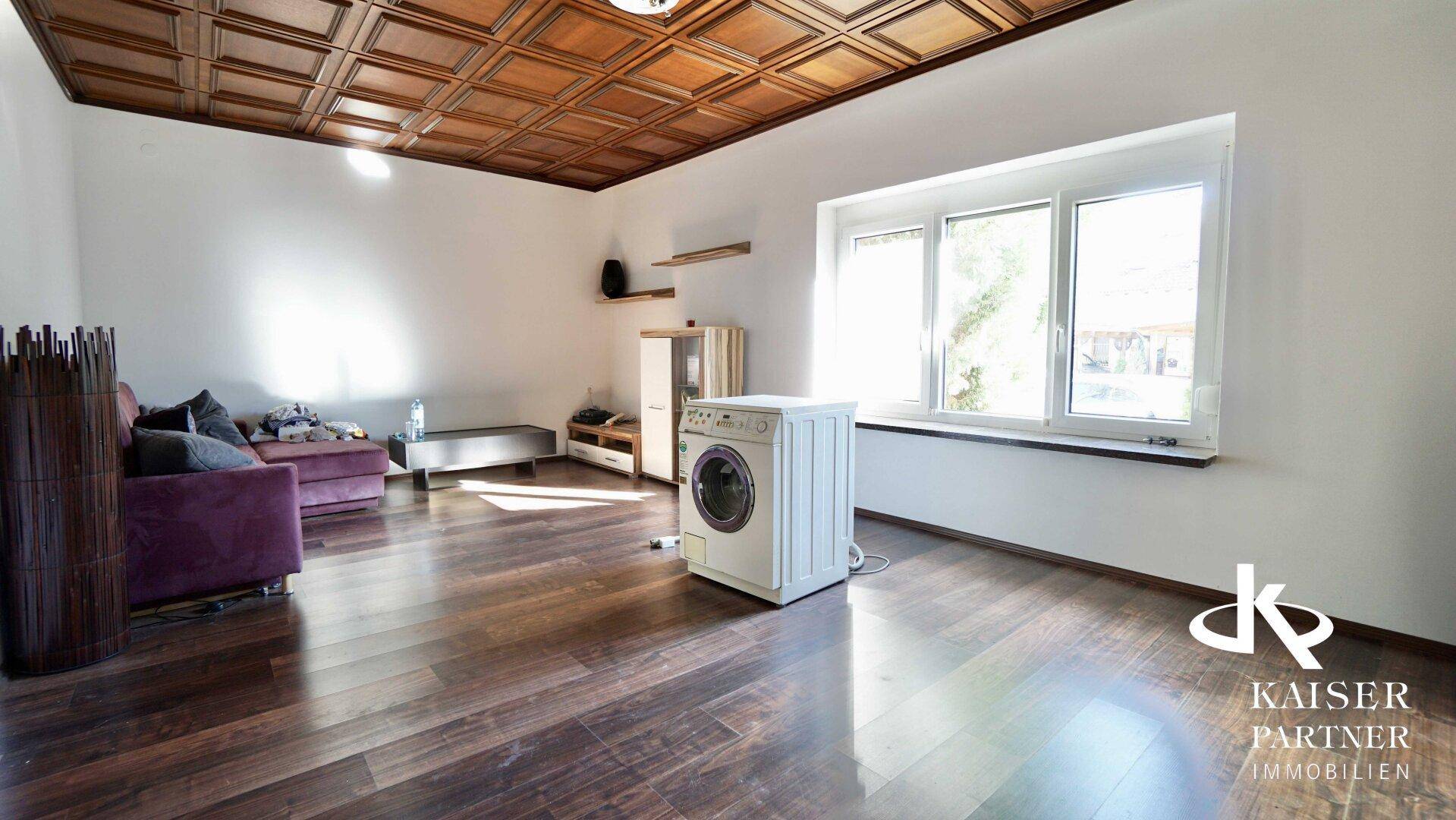 Geräumiges Zimmer mit dunklem Holzboden, Kassettendecke und großem Fenster, inklusive Waschmaschine.