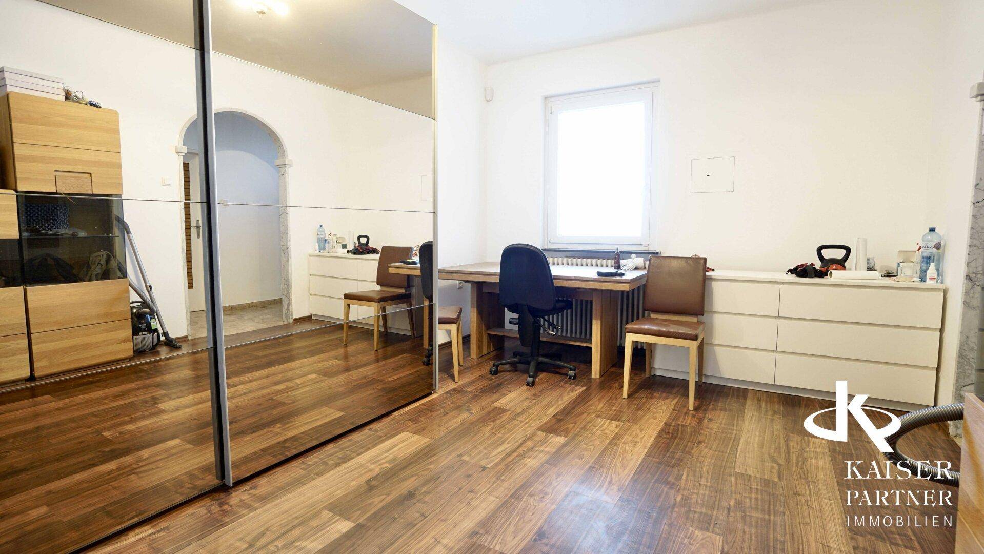 Helles Arbeitszimmer mit dunklem Holzboden, großem Spiegelschrank und einem Fenster für Tageslicht.