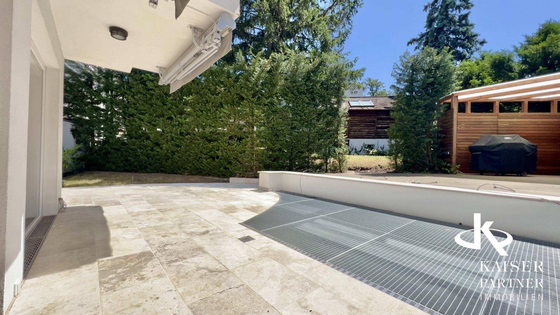 Geräumige Steinterrasse mit Fliesenboden, umgeben von einer hohen Hecke und einem Grillbereich.