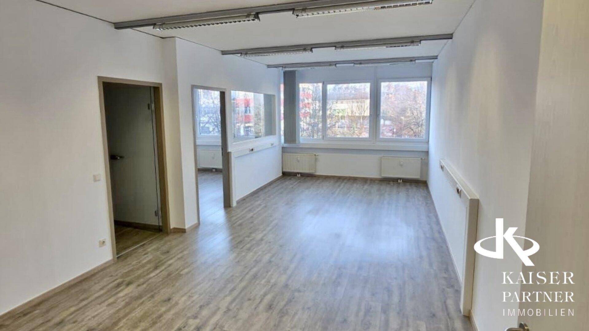 Geräumiges, leeres Büro mit großen Fenstern, hellem Laminatboden und Blick ins Grüne.