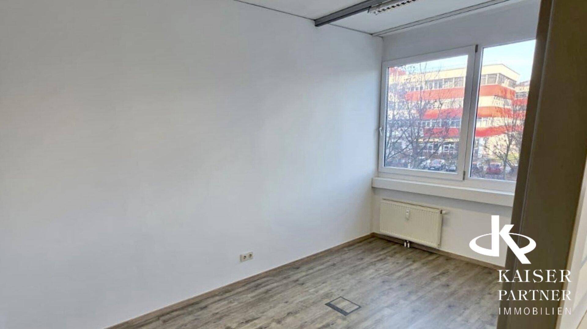 Kleineres, helles Büro mit einem großen Fenster, Laminatboden und Heizkörper unter dem Fenster.