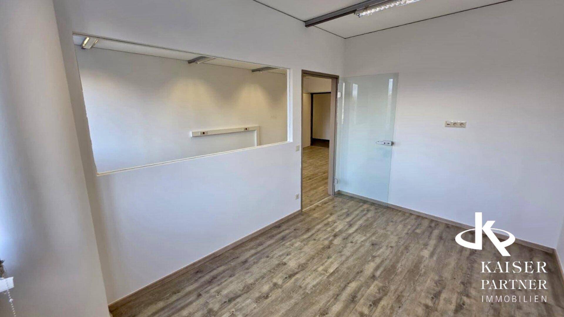 Leeres Büro mit Laminatboden, einer Glastür und einer halbhohen Trennwand zu einem Nebenraum.