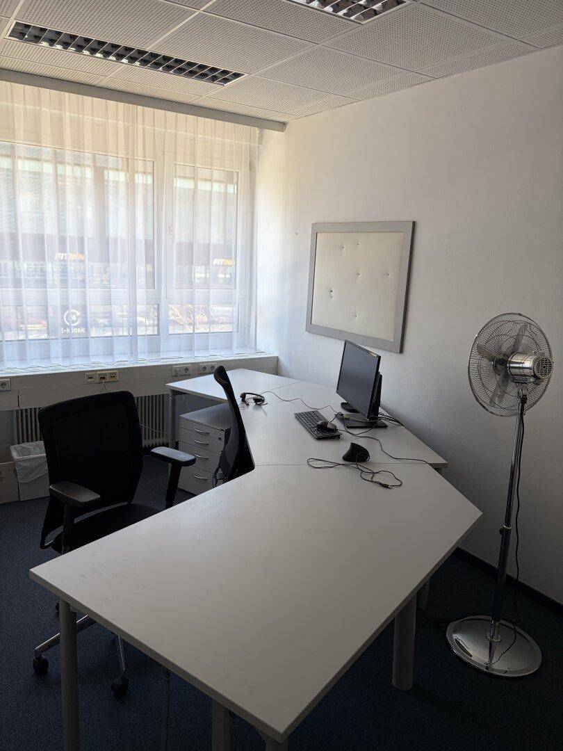 Funktionales Büro mit einem L-förmigen Schreibtisch, Computer und einem Ventilator, gut beleuchtet durch ein Fenster.