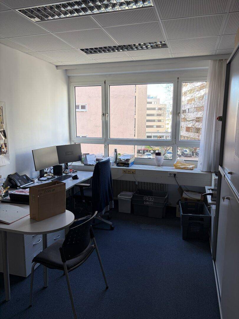 Helles Büro mit zwei Arbeitsplätzen, Computermonitoren und einem Fenster mit Blick auf andere Gebäude.