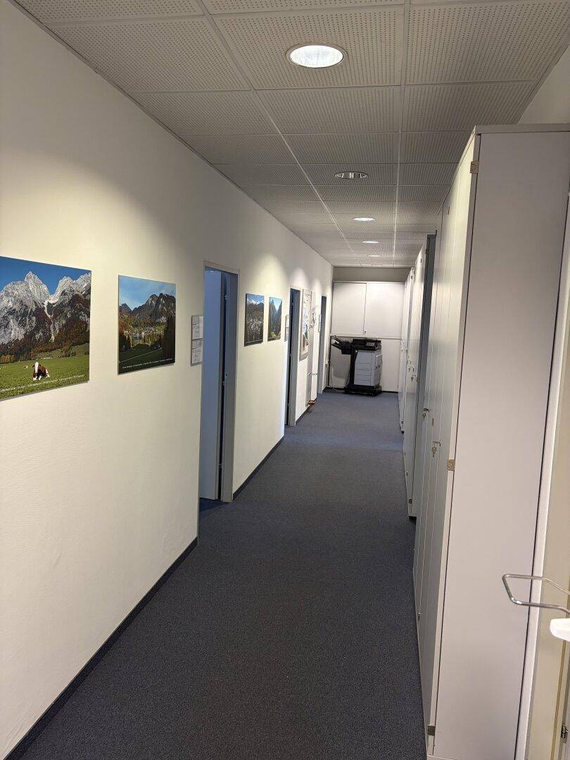Langer Bürogang mit Bildern an der Wand, Einbauschränken und Teppichboden, der zu weiteren Räumen führt.