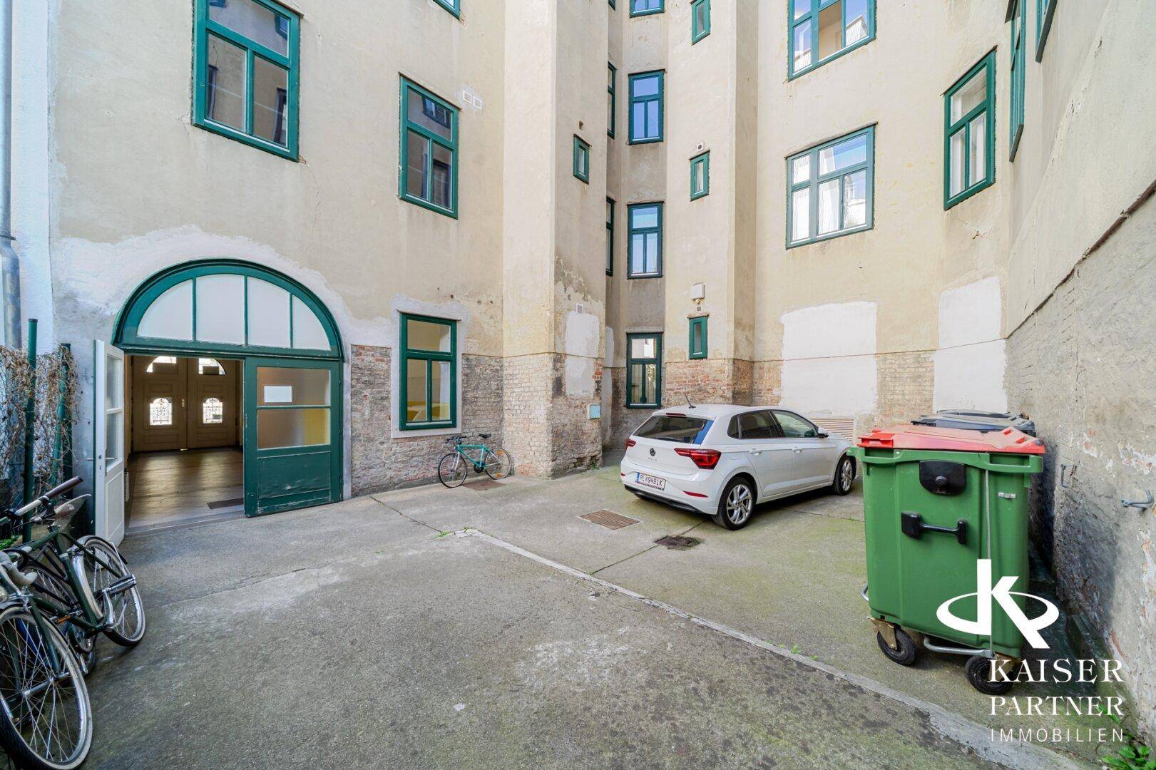 Innenhof eines Gebäudes mit Parkplätzen, Fahrrädern und Mülltonnen, umgeben von Fassaden.