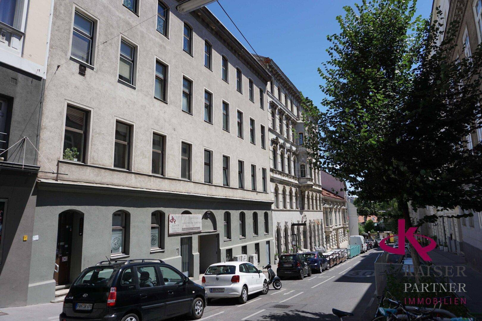 Mehrstöckiges Wohngebäude an einer städtischen Straße mit parkenden Autos und klarem Himmel.