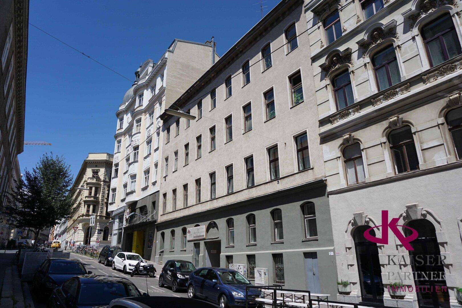Städtische Straßenszene mit historischen Gebäudefassaden und parkenden Fahrzeugen unter blauem Himmel.