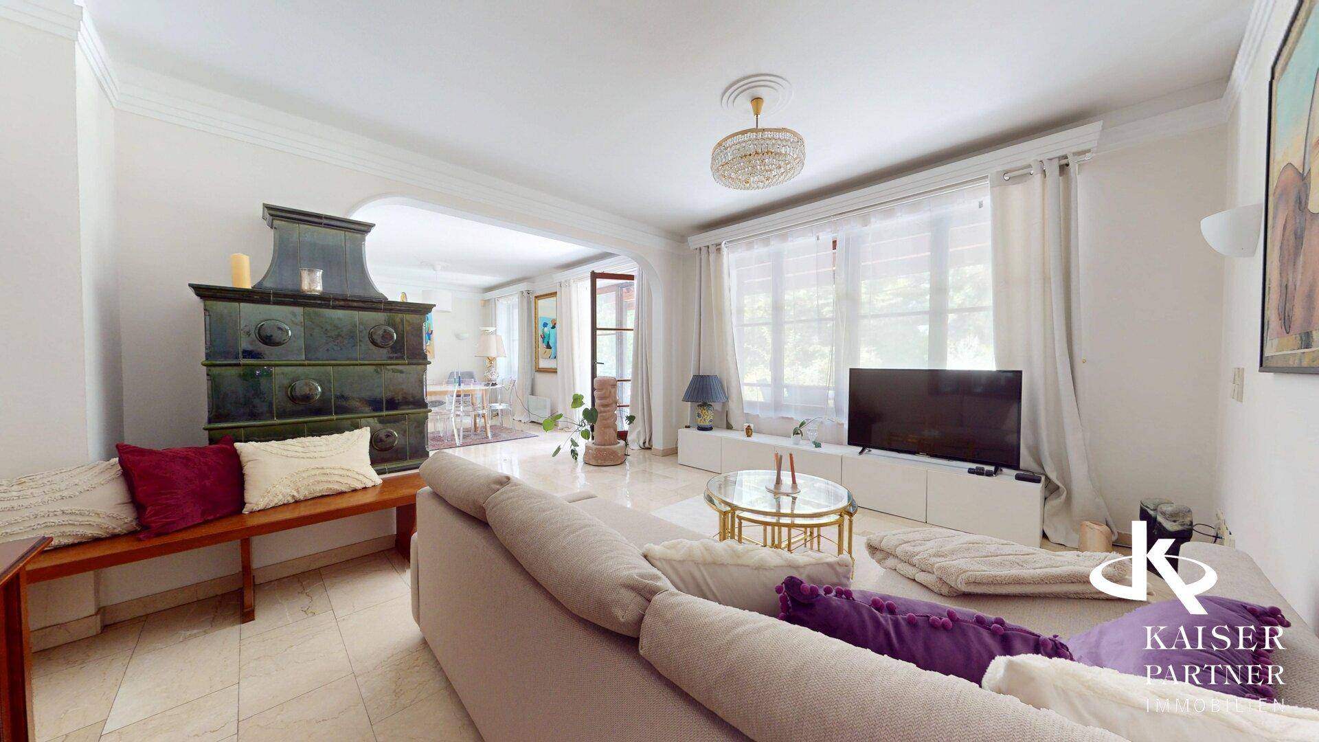 Gemütliches Wohnzimmer mit hellem Sofa, Fernseher und traditionellem Kachelofen.