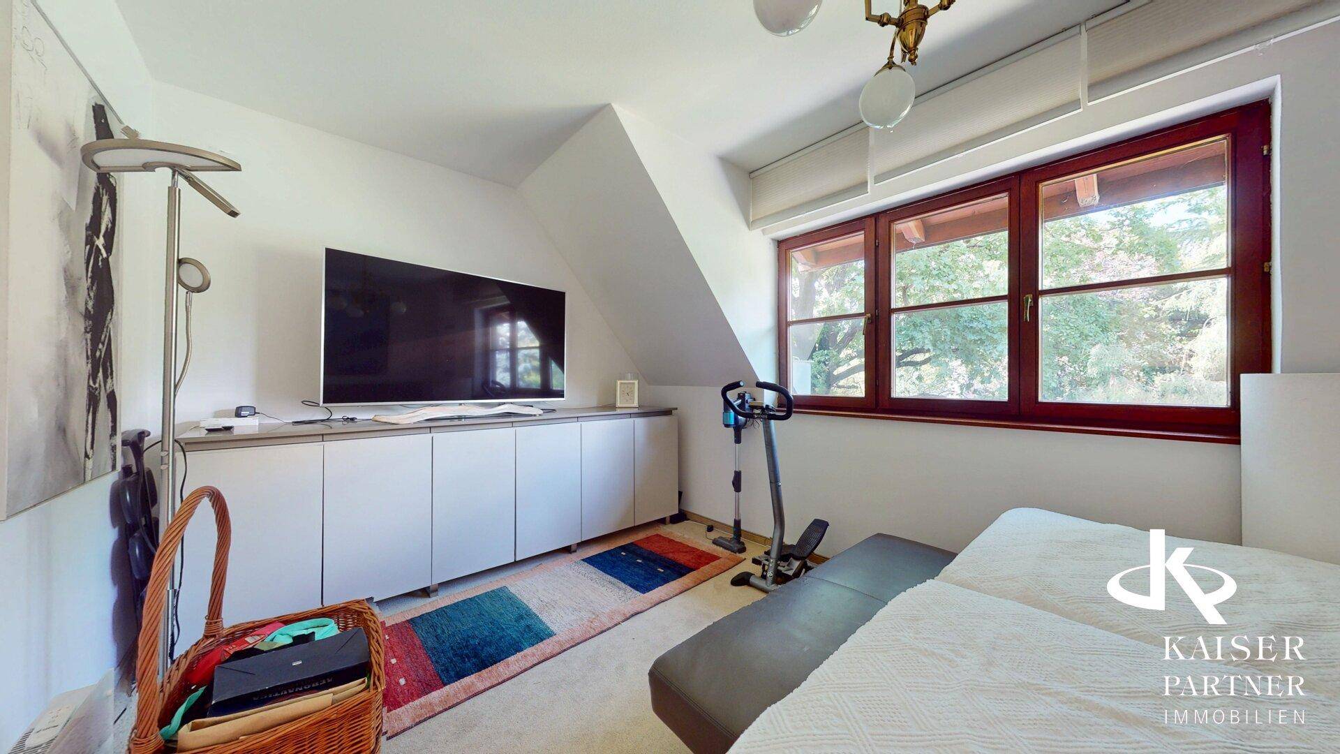 Schlafzimmer im Dachgeschoss mit großem Fernseher, Fitnessgerät und Blick ins Grüne.