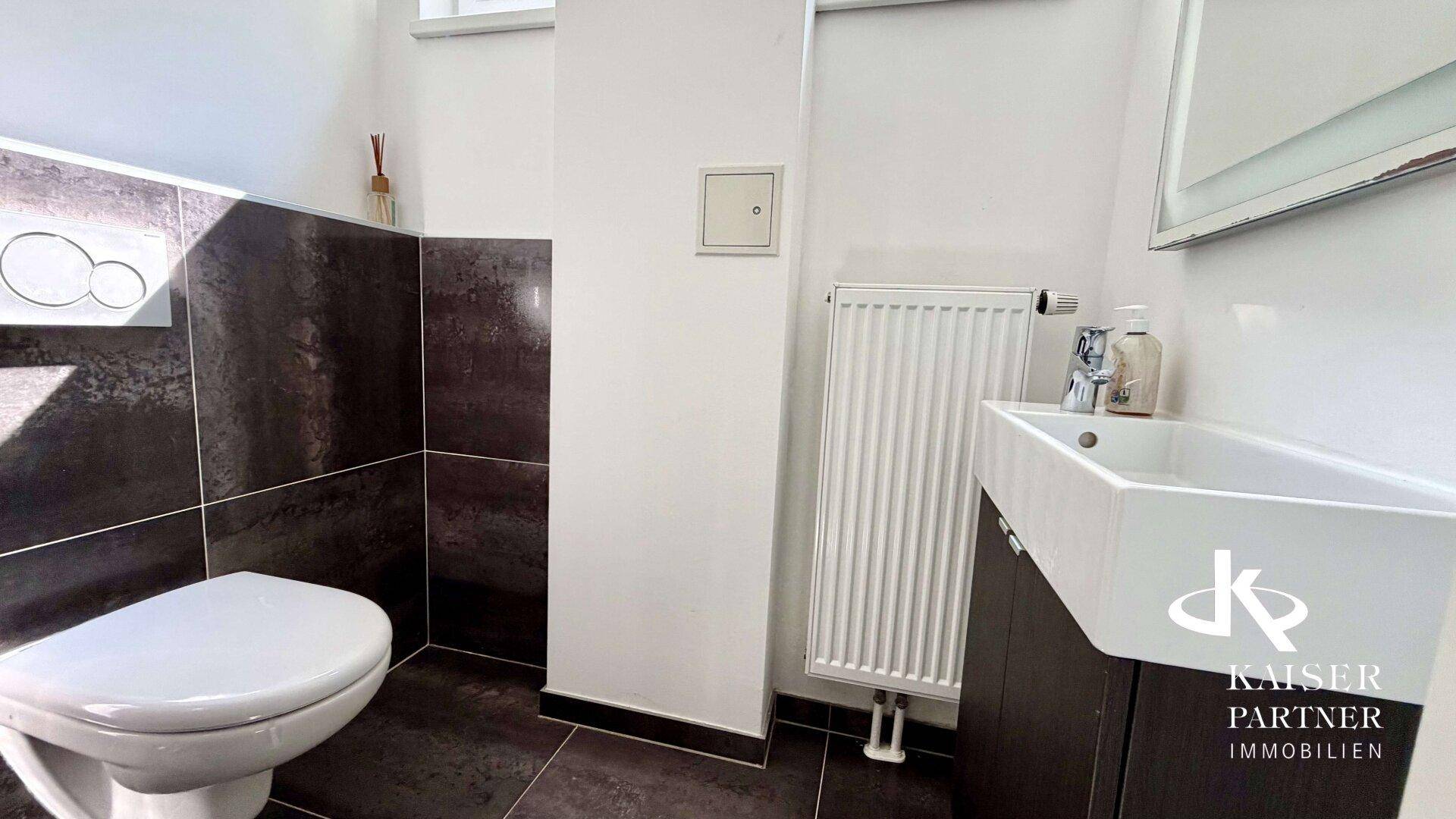 Modernes Gäste-WC mit dunklen Fliesen, wandhängender Toilette und kleinem Waschbecken mit Unterschrank.