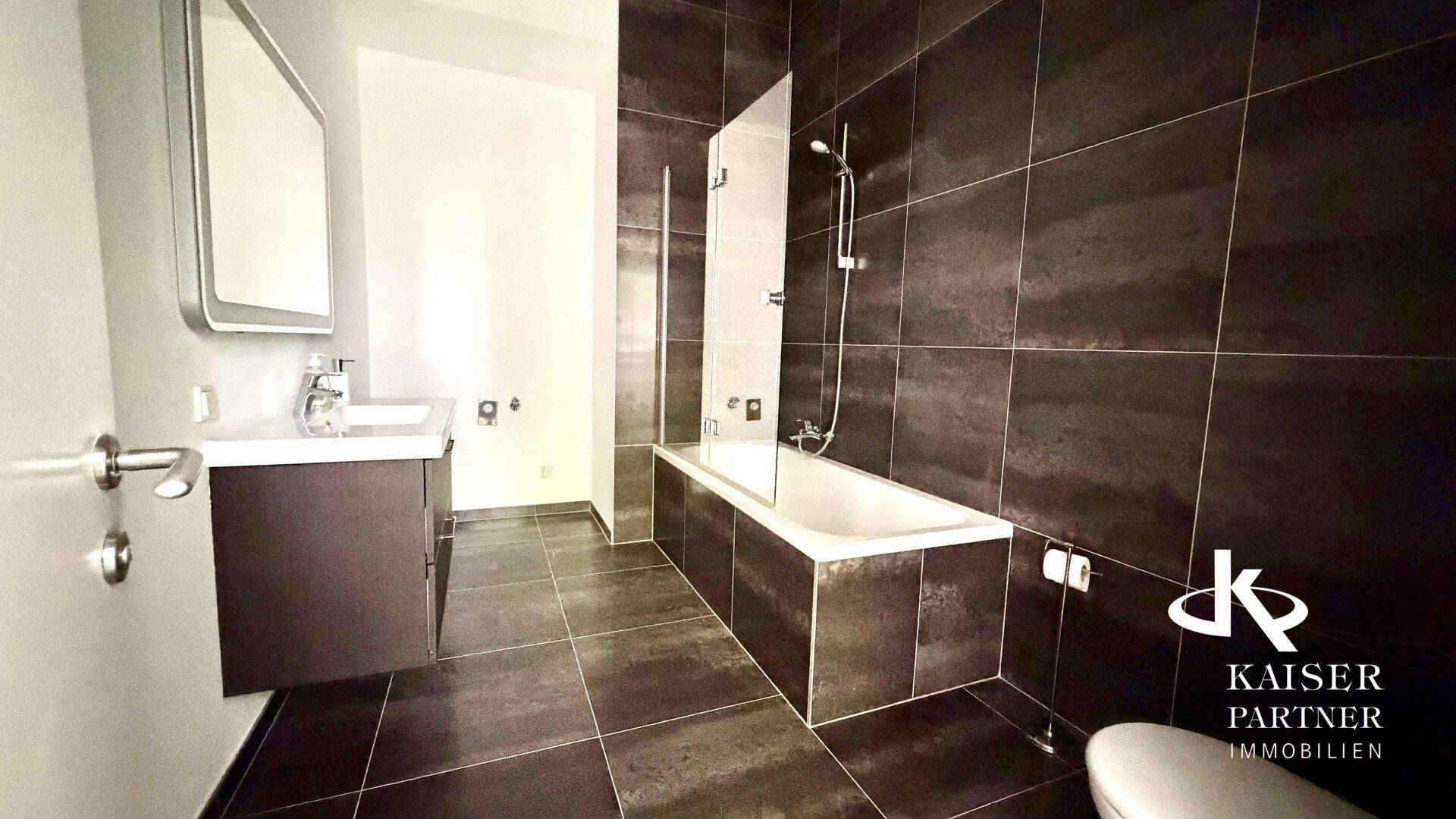 Modernes Badezimmer mit dunklen Fliesen, Badewanne mit Duschabtrennung und Waschbecken mit Spiegel.