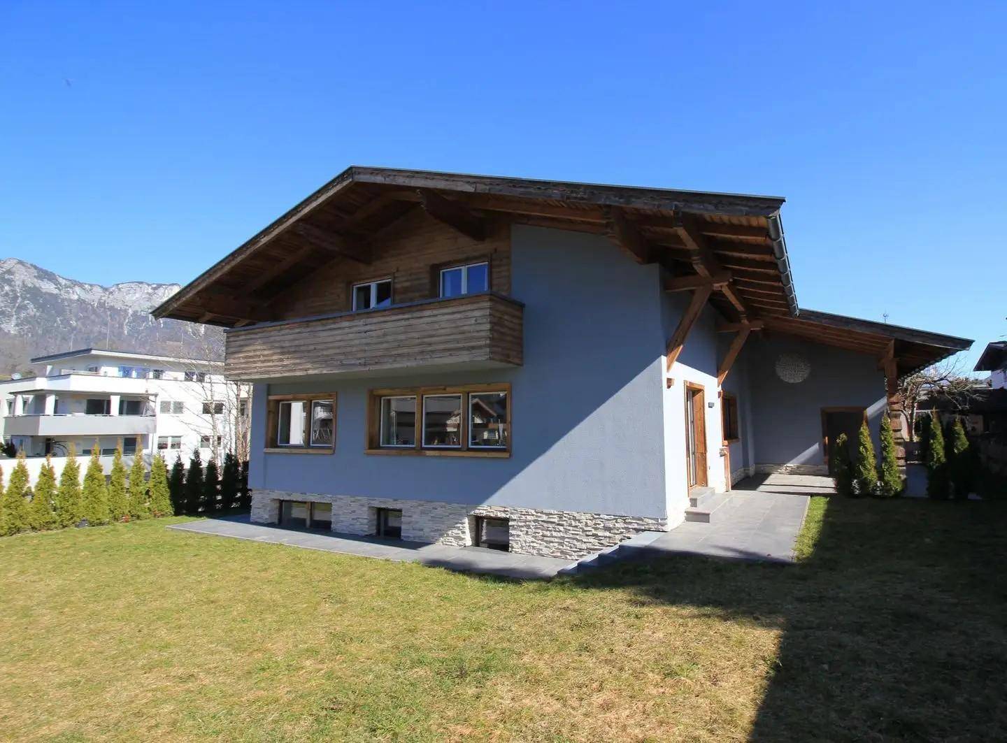 Modernes Haus mit Holzdetails, Balkon und gepflegtem Rasen vor Bergkulisse.