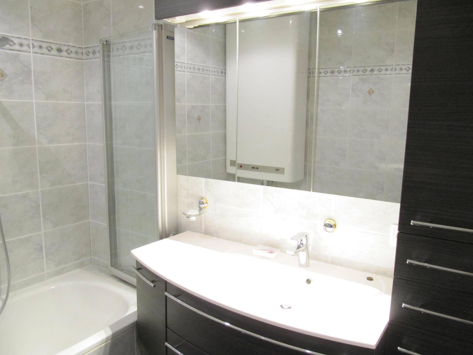 Badezimmer mit Waschbecken, Spiegelschrank und Badewanne, in hellen Grautönen gefliest.