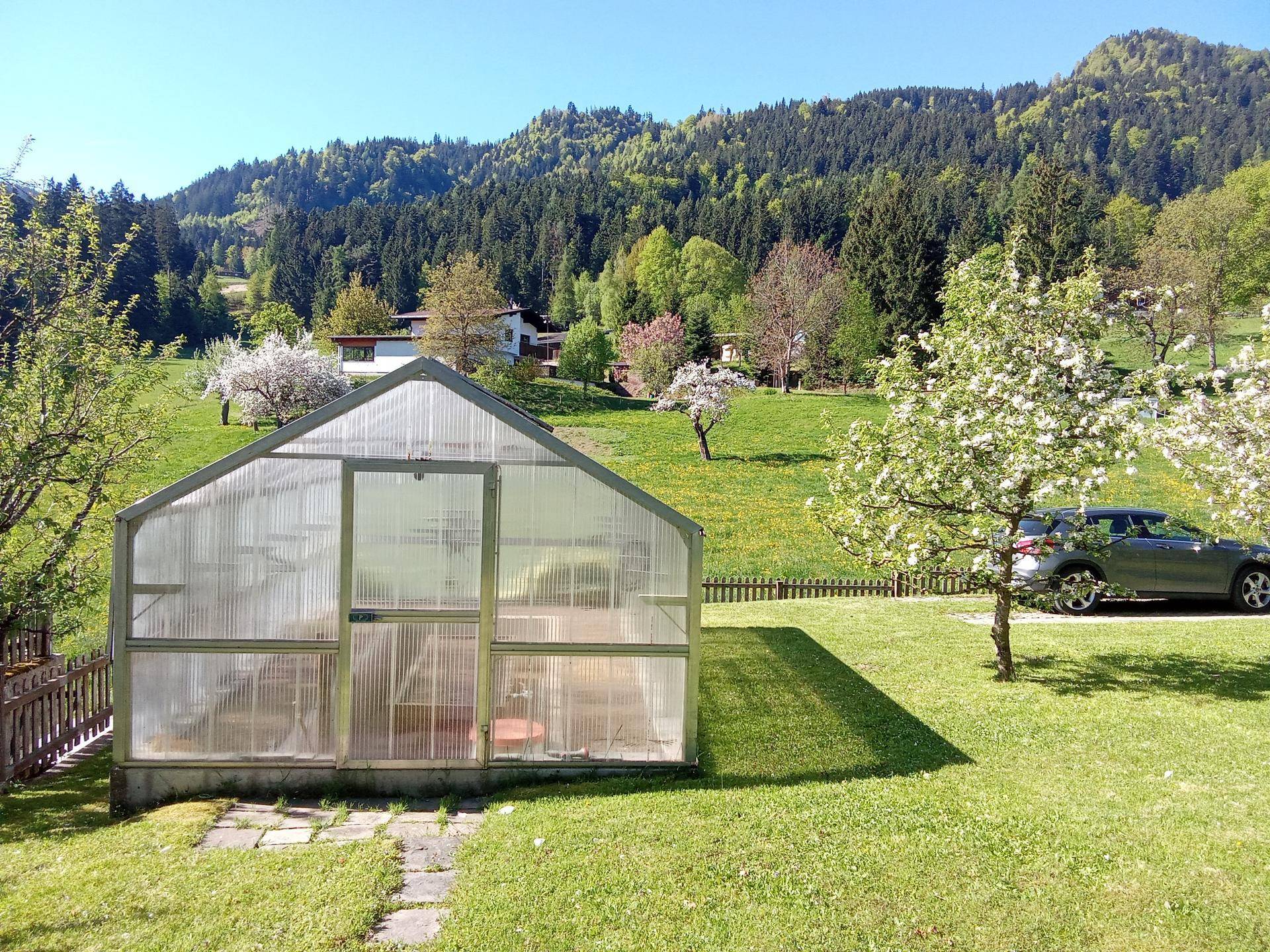 Gepflegter Gartenbereich mit Gewächshaus und blühenden Obstbäumen vor bewaldeten Hügeln.