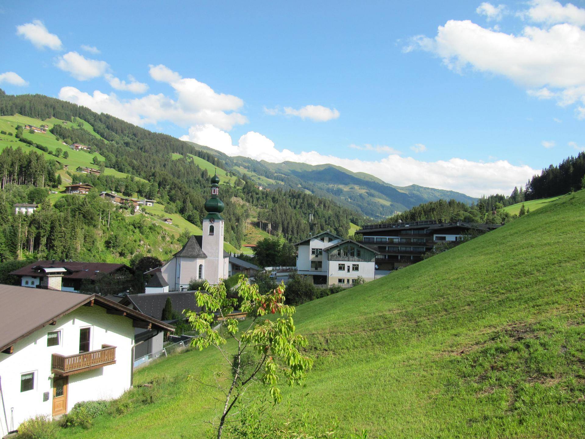 Panoramablick auf ein idyllisches Dorf in den Alpen mit einer markanten Kirche, umgeben von grünen Hügeln und Bergen unter blauem Himmel.
