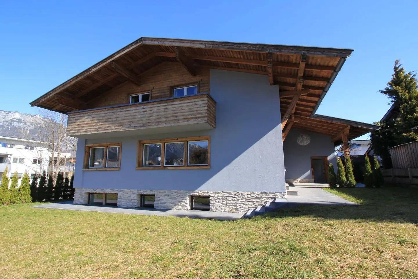 Charmantes Haus mit Holzbalkon, grauer Fassade und gepflegtem Gartenbereich.