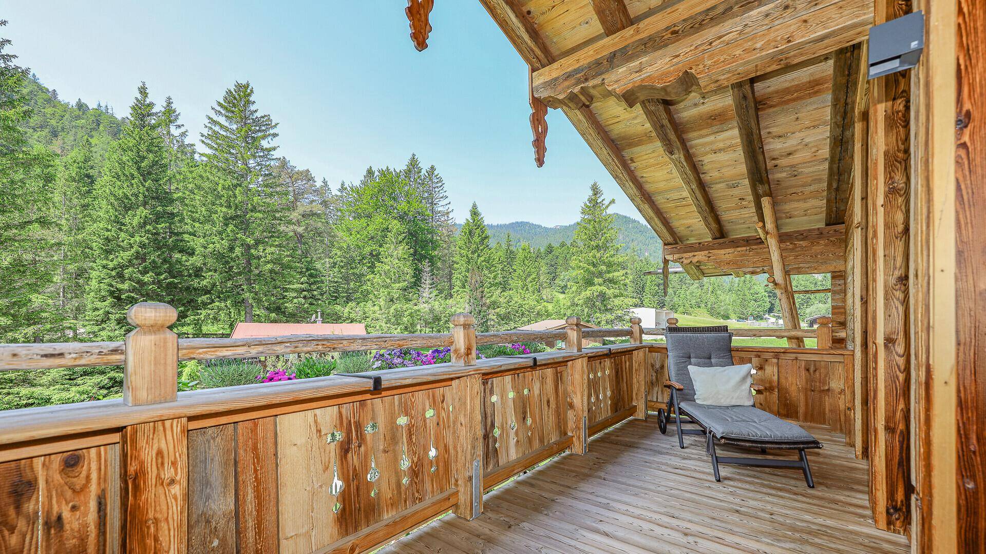 Gemütlicher Balkon mit Holzgeländer, Liegestuhl und Blick auf den umliegenden Wald und die Berge.