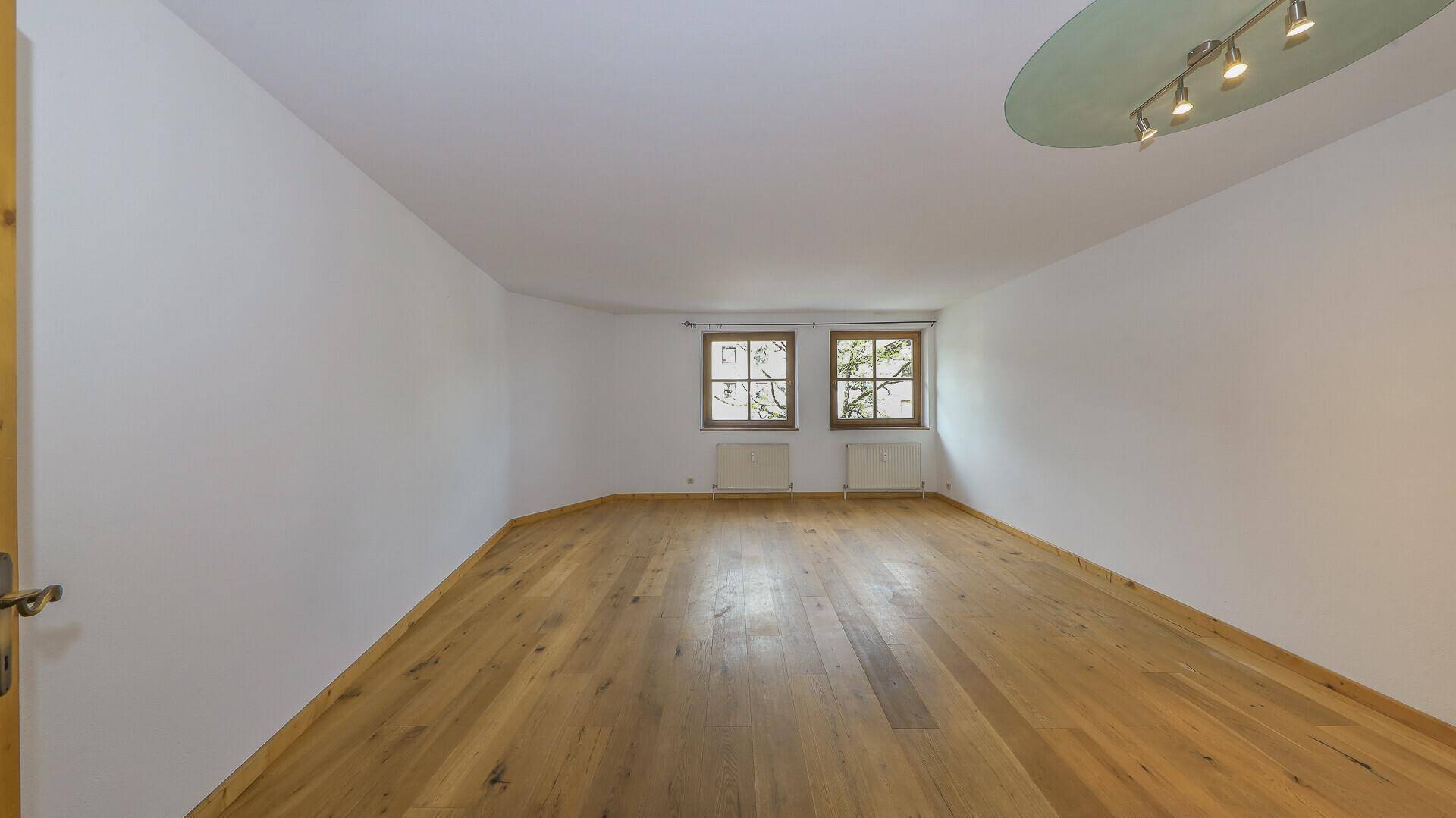 Helles, leeres Zimmer mit Holzboden und zwei Fenstern, ideal für ein Wohn- oder Schlafzimmer.