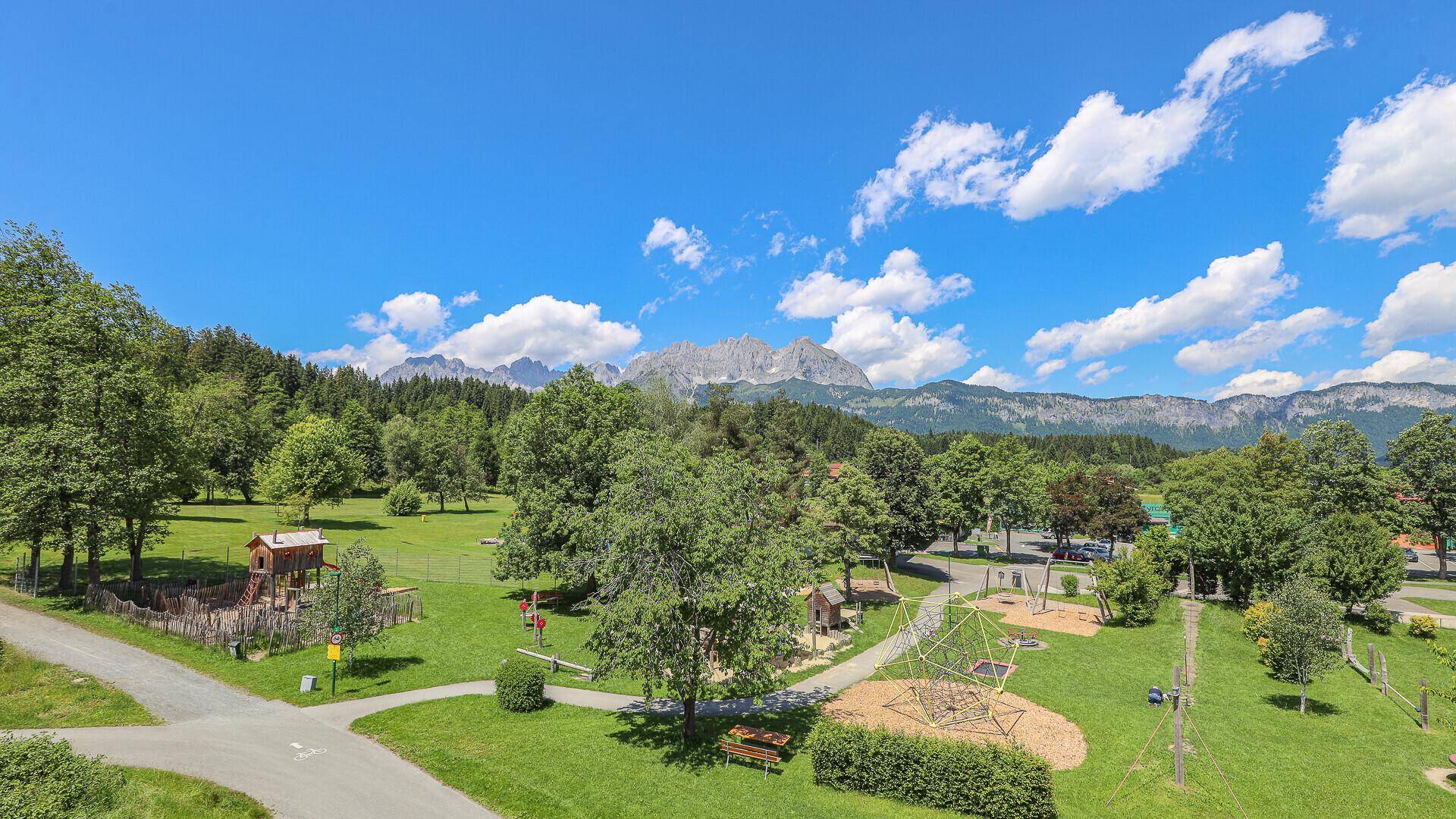 Weitläufige Grünfläche mit Spielplatz, Bäumen und beeindruckendem Bergpanorama im Hintergrund.