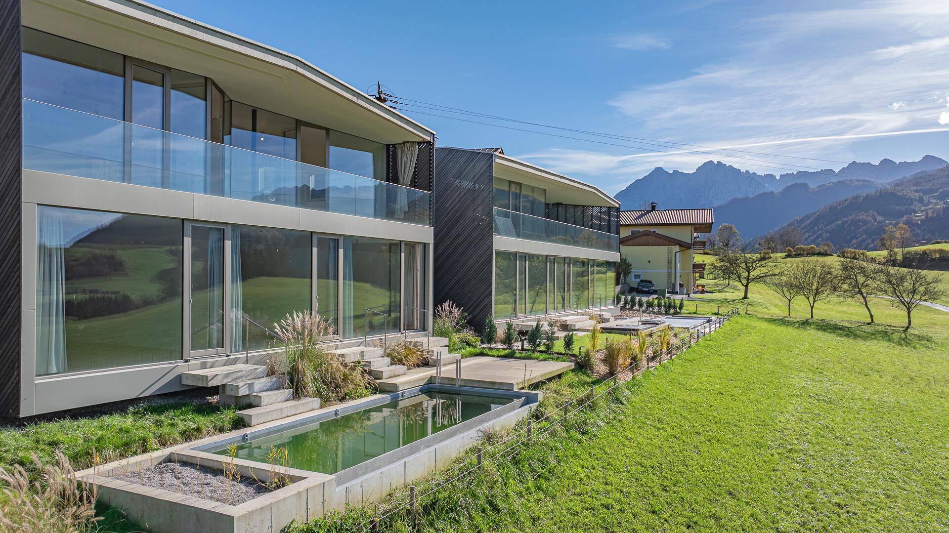 Moderne Architektur mit großen Glasfronten, Balkonen und einem kleinen Pool, umgeben von grünen Wiesen und Bergblick.
