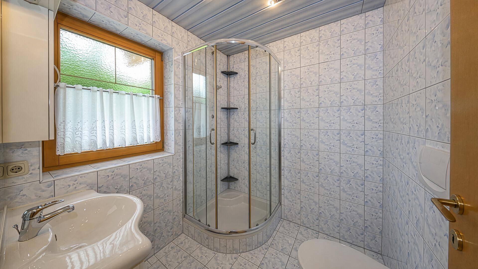 Helles Badezimmer mit Duschkabine, Waschbecken und Fenster mit Blick ins Grüne.