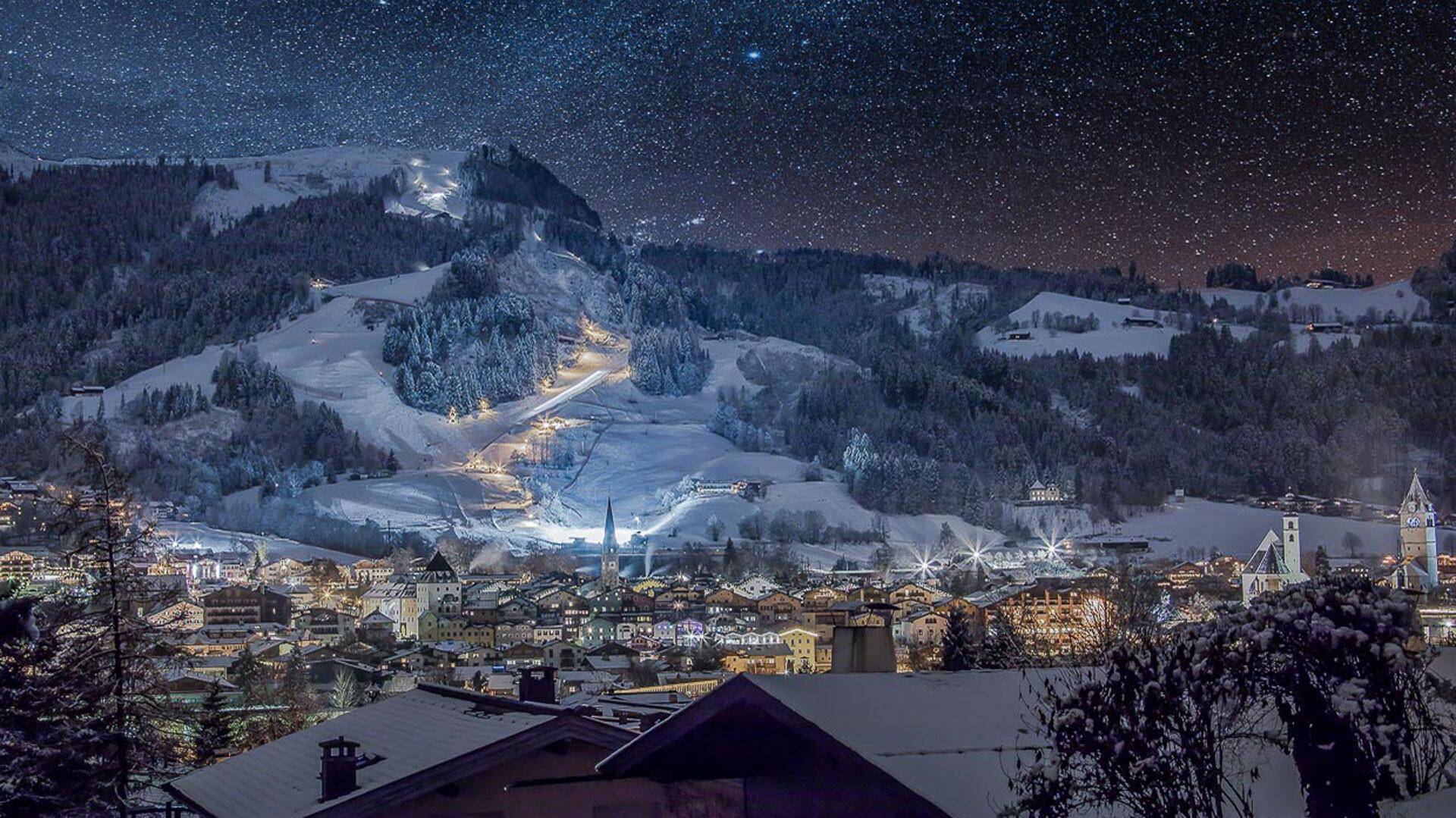 Nachtaufnahme einer verschneiten Stadt und Berglandschaft unter einem klaren Sternenhimmel.