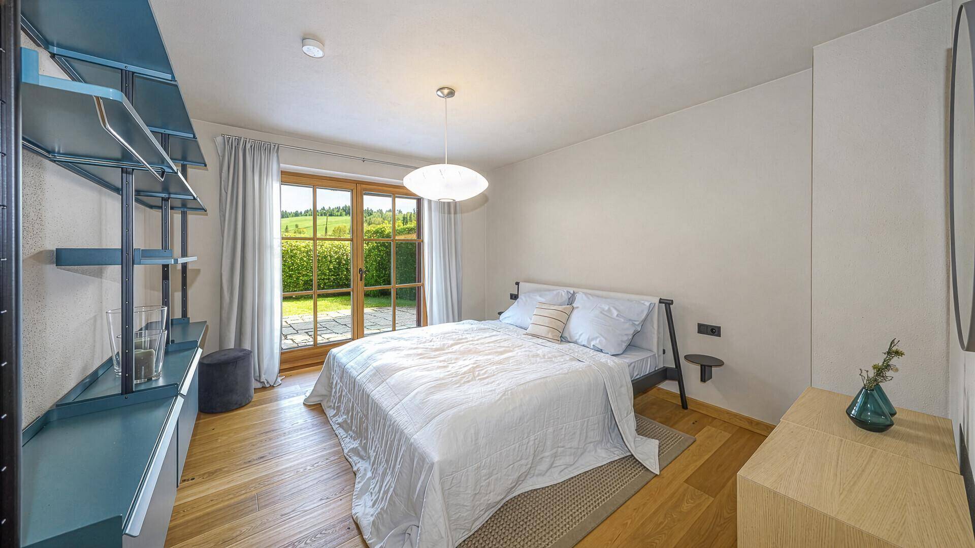Helles Schlafzimmer mit Holzboden, Doppelbett und Blick ins Grüne durch große Fenster.