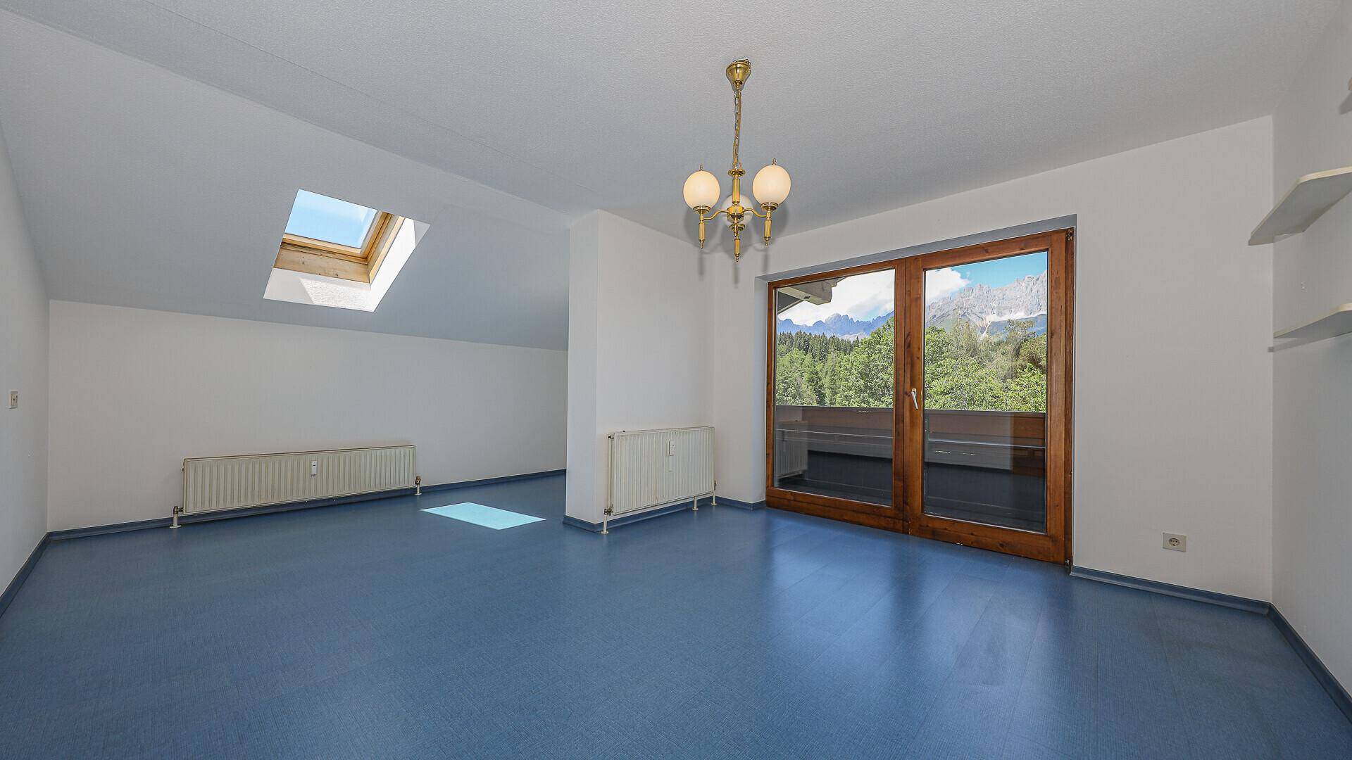 Leeres Wohnzimmer mit blauem Boden, Dachfenster und Zugang zum Balkon mit Bergblick.