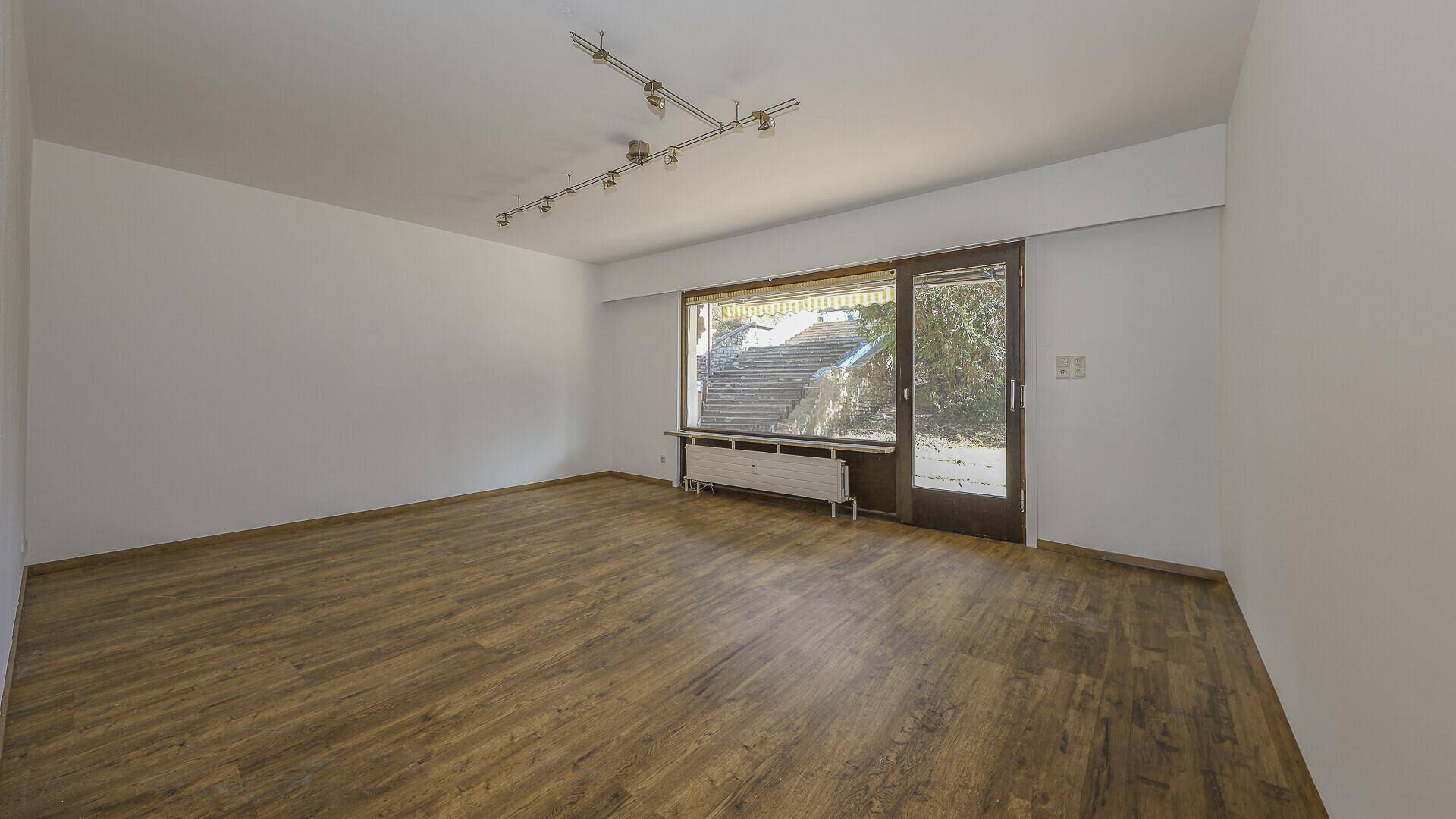 Heller, leerer Wohnraum mit Holzboden und großem Fenster, das viel Tageslicht hereinlässt.