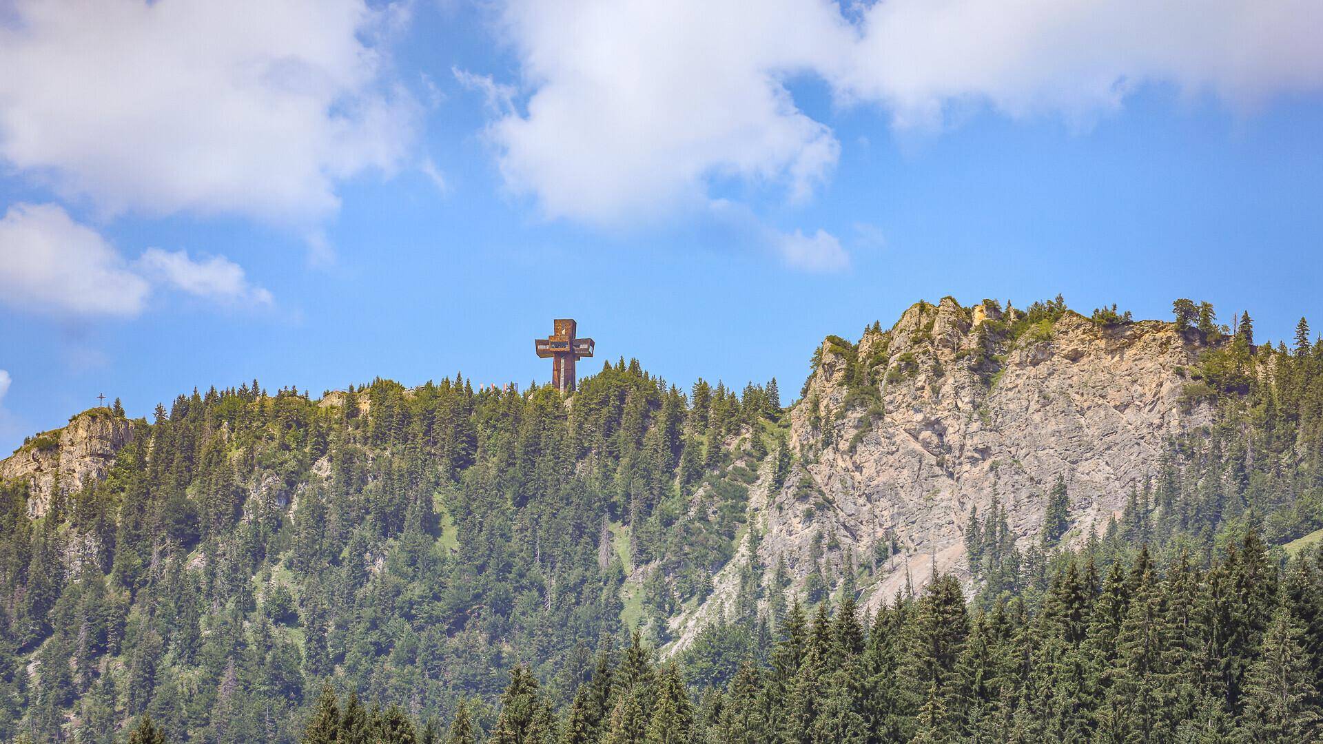 Gipfelkreuz auf einem bewaldeten Berg mit felsigen Klippen unter einem klaren blauen Himmel.