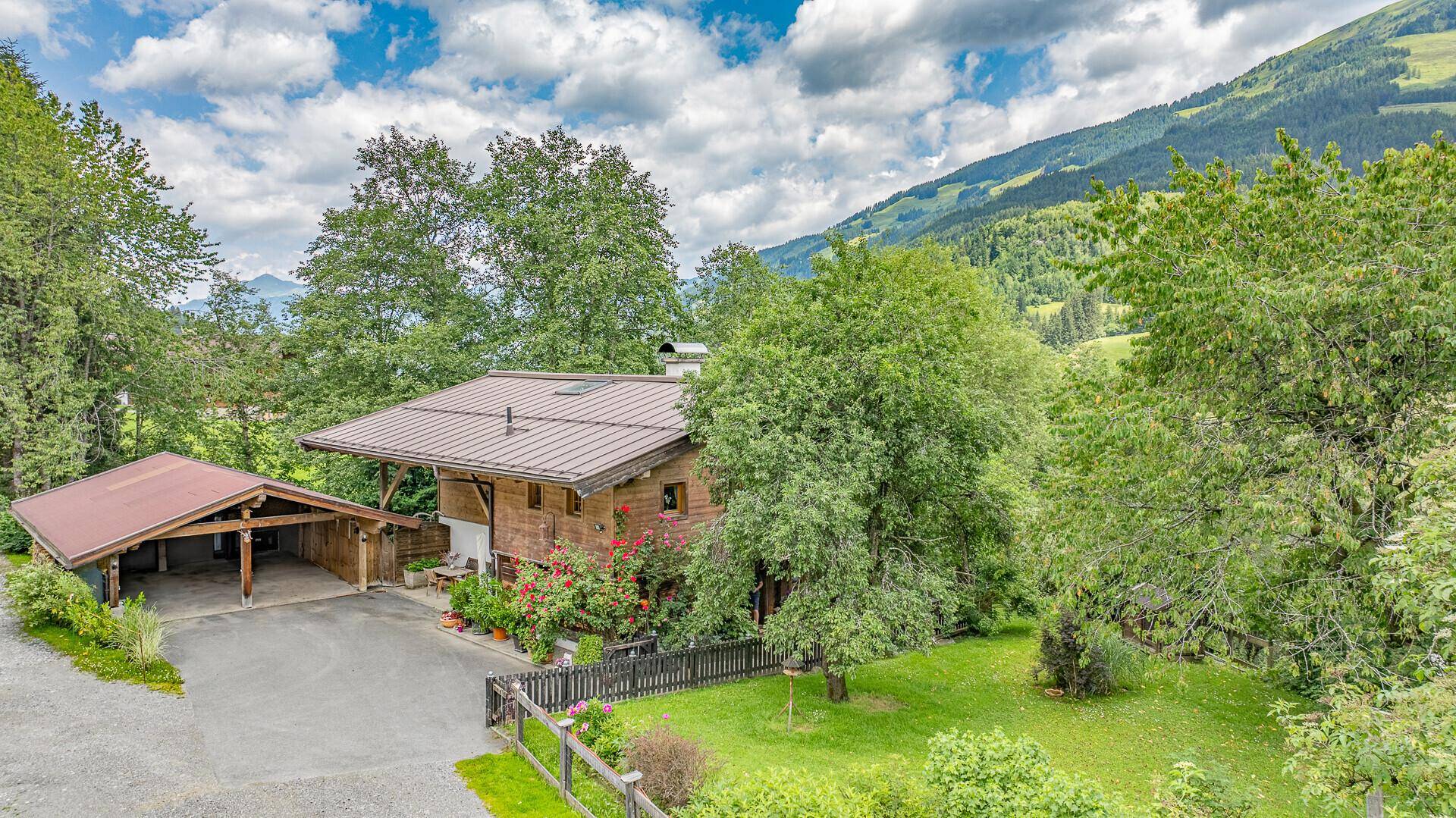 Idyllisches Holzhaus mit Carport und großem Garten, umgeben von grüner Natur und Bergen.