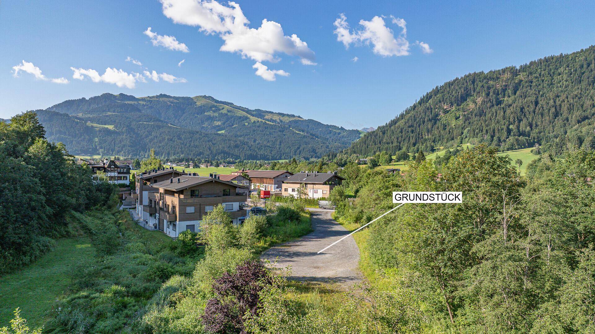 Panoramablick auf das Grundstück mit umliegenden Häusern und grünen Bergen unter blauem Himmel.