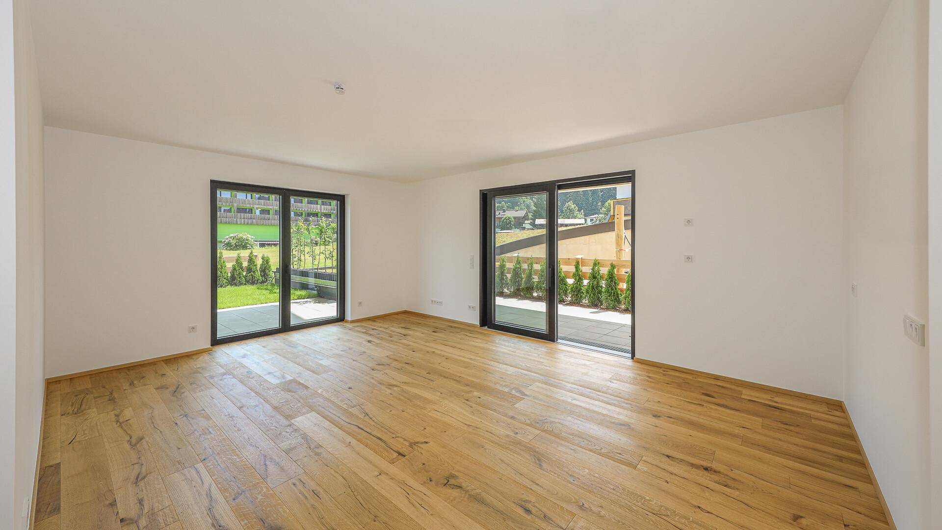 Geräumiges, leeres Zimmer mit Holzboden und großen Fenstern zum Gartenbereich.