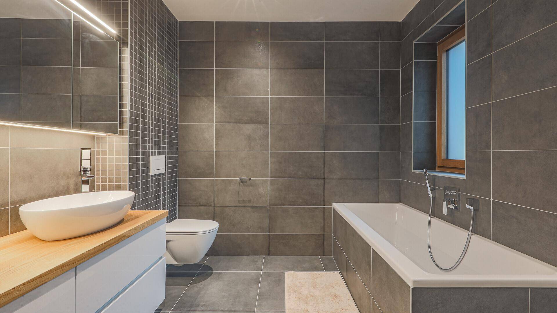 Modernes Badezimmer mit großen grauen Fliesen, Badewanne, Waschtisch und Wand-WC.