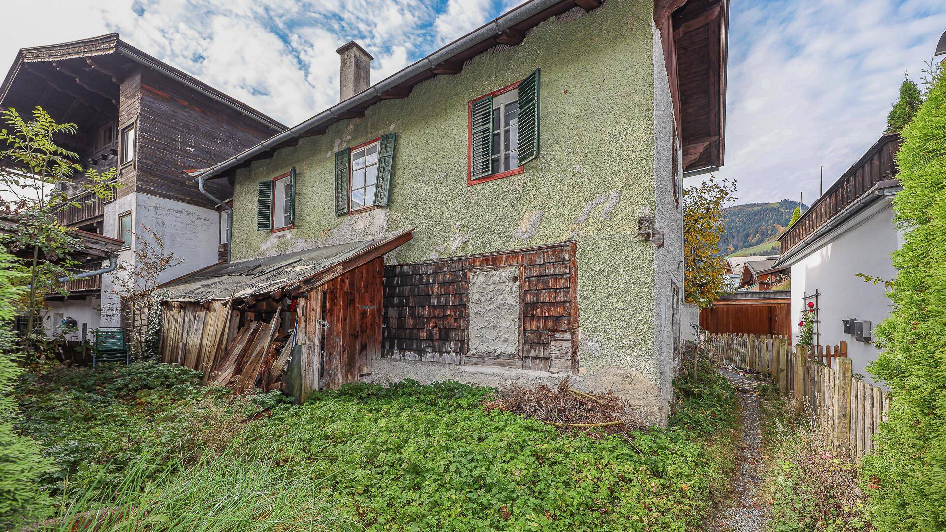 Rückseite des alten Hauses mit grünem Putz, verfallenem Schuppen und verwildertem Gartenbereich.