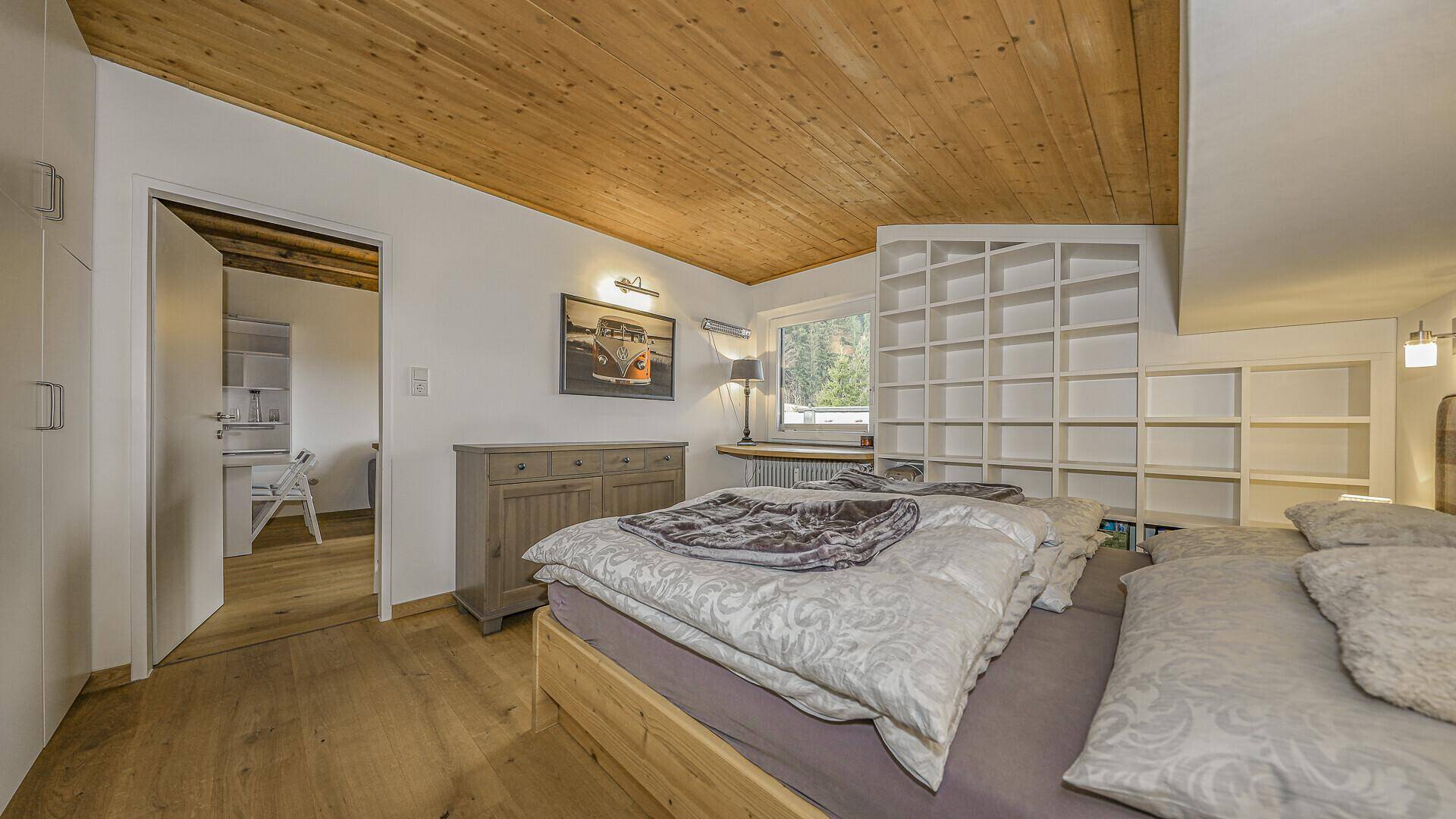Helles Schlafzimmer mit Doppelbett, offenen Regalen und Holzdecke, mit Zugang zu einem weiteren Raum.
