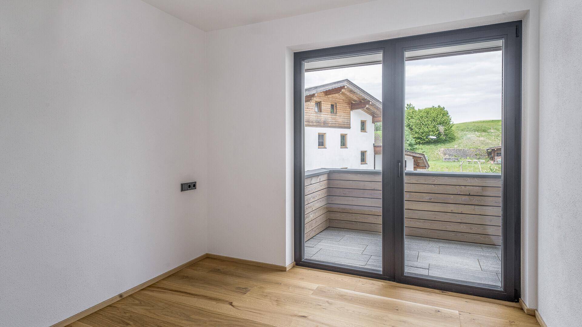 Helles Zimmer mit Holzboden und großen Fenstern, die Zugang zu einem Balkon bieten.