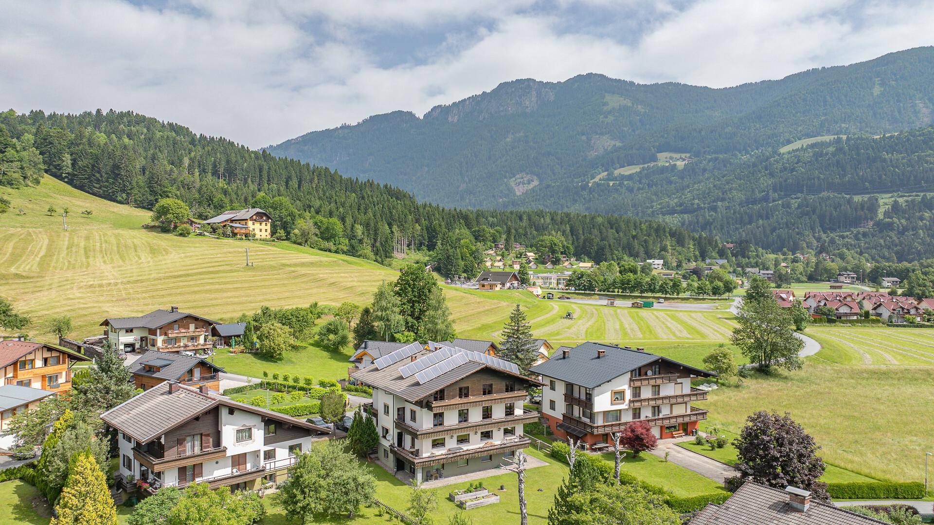 Weitläufige Luftaufnahme eines Dorfes eingebettet in eine grüne Hügellandschaft mit majestätischen Bergen im Hintergrund.