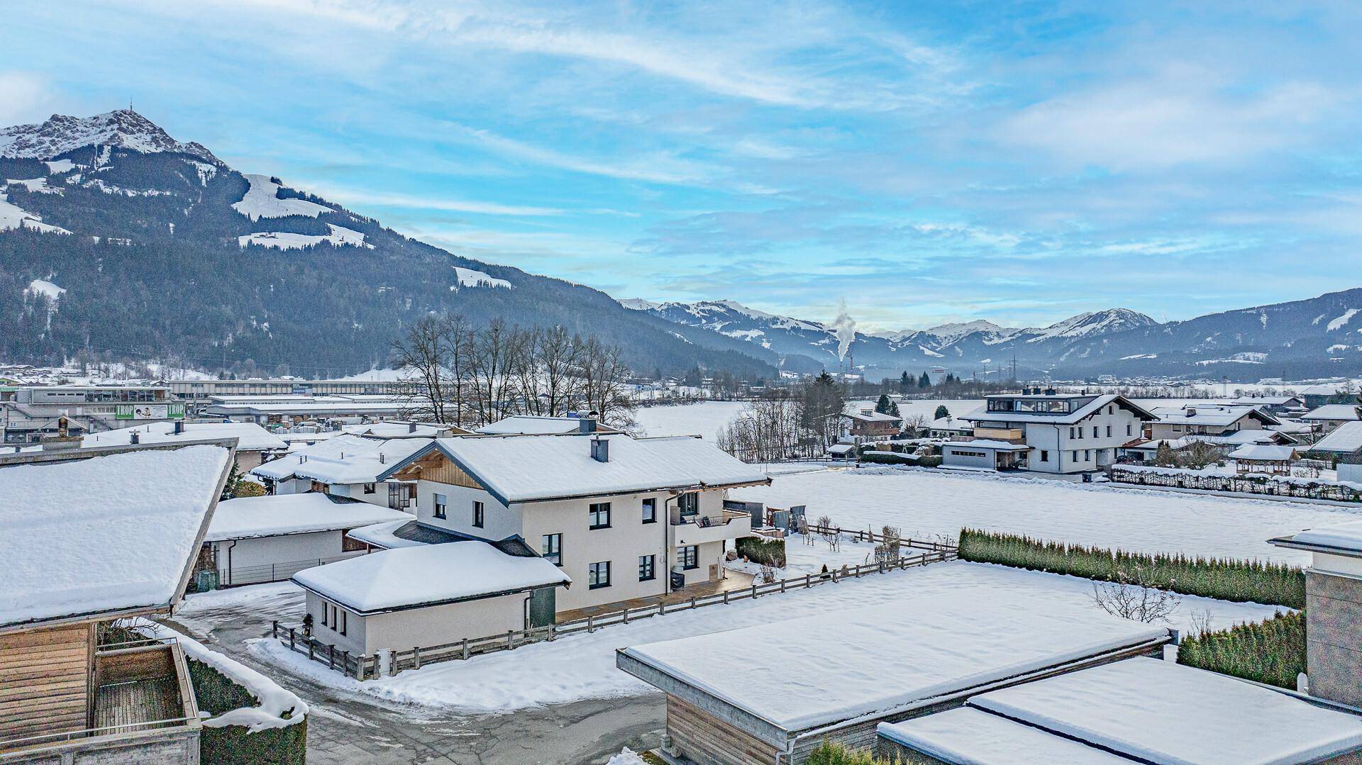 Luftaufnahme einer Siedlung in den Bergen mit verschneiten Dächern und weitem Blick ins Tal.
