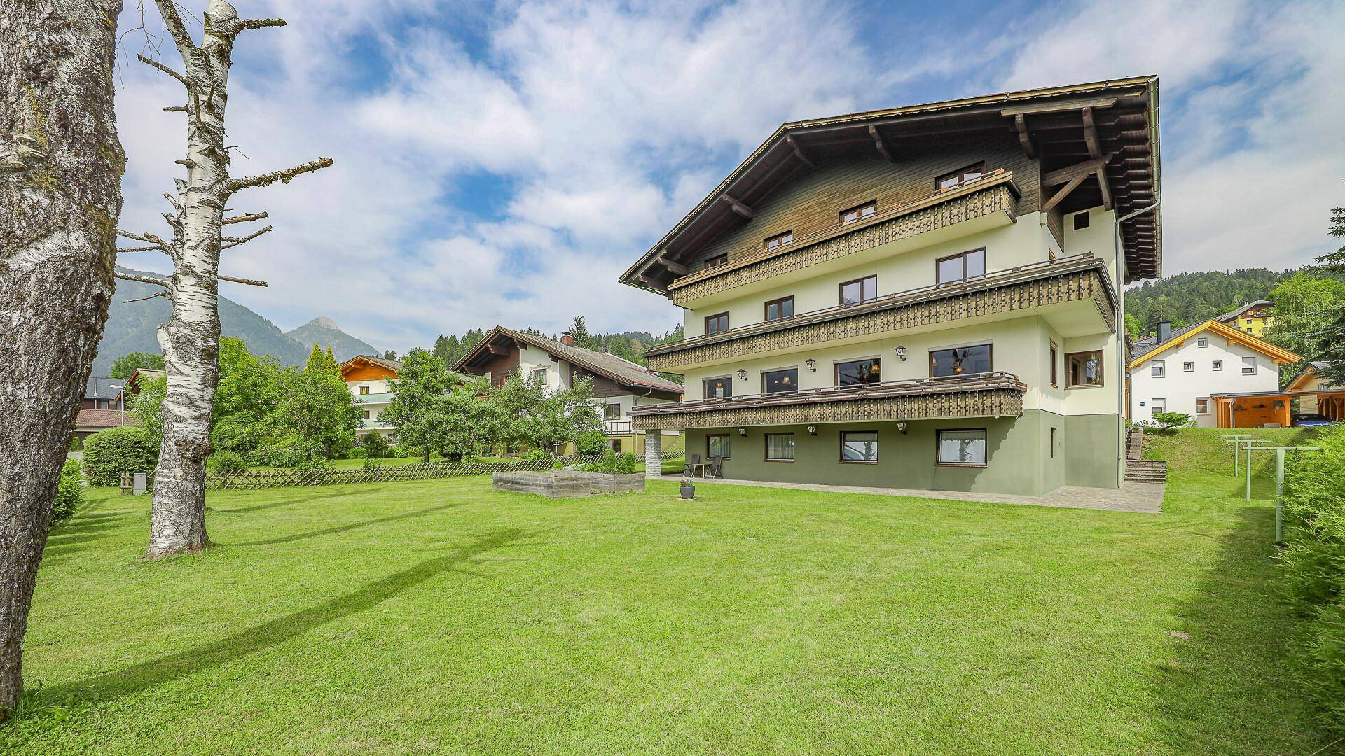 Ansicht des Gebäudes vom Garten aus, mit weißer Fassade, grüner Basis und mehreren Balkonen.