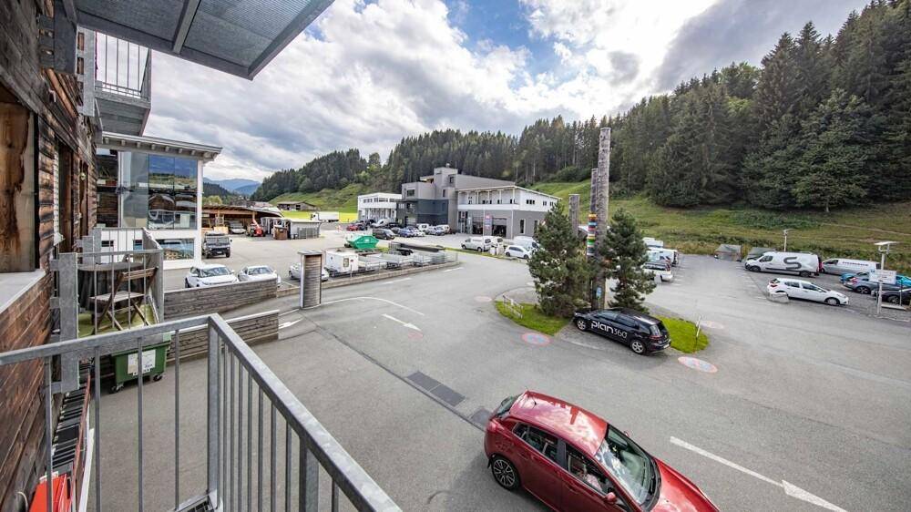 Blick vom Balkon auf einen Parkplatz, umliegende Gebäude und bewaldete Hügel unter bewölktem Himmel.