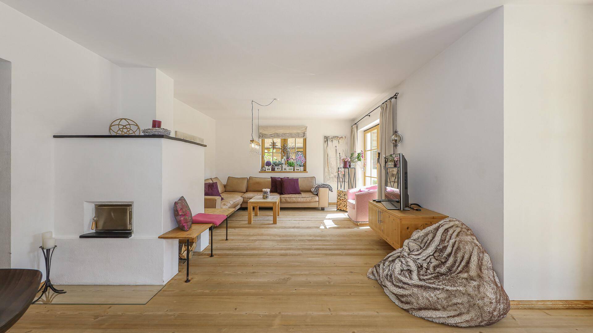 Geräumiges Wohnzimmer mit Kachelofen, Holzboden und Blick auf den Gartenbereich.