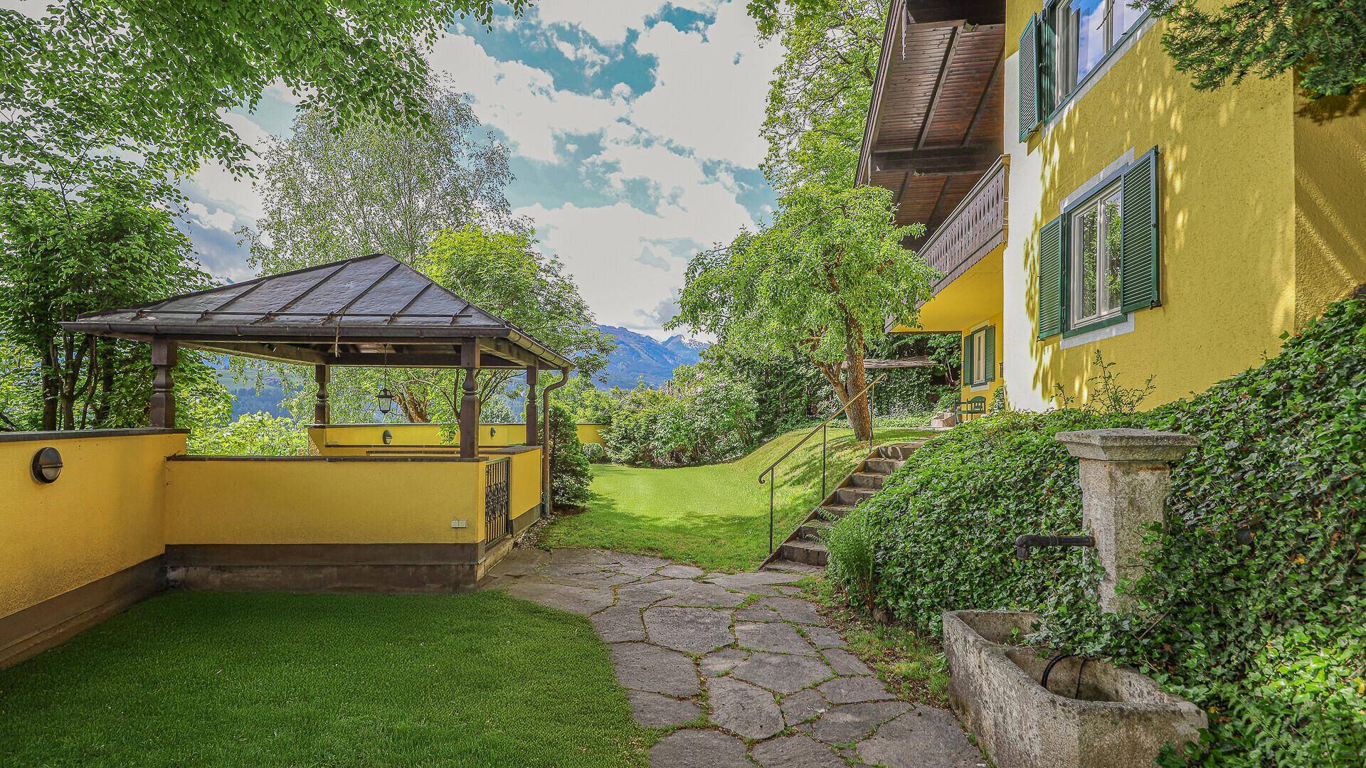 Seitlicher Gartenweg mit Steinplatten, historischem Brunnen und Blick auf das gelbe Haus und die Berge.