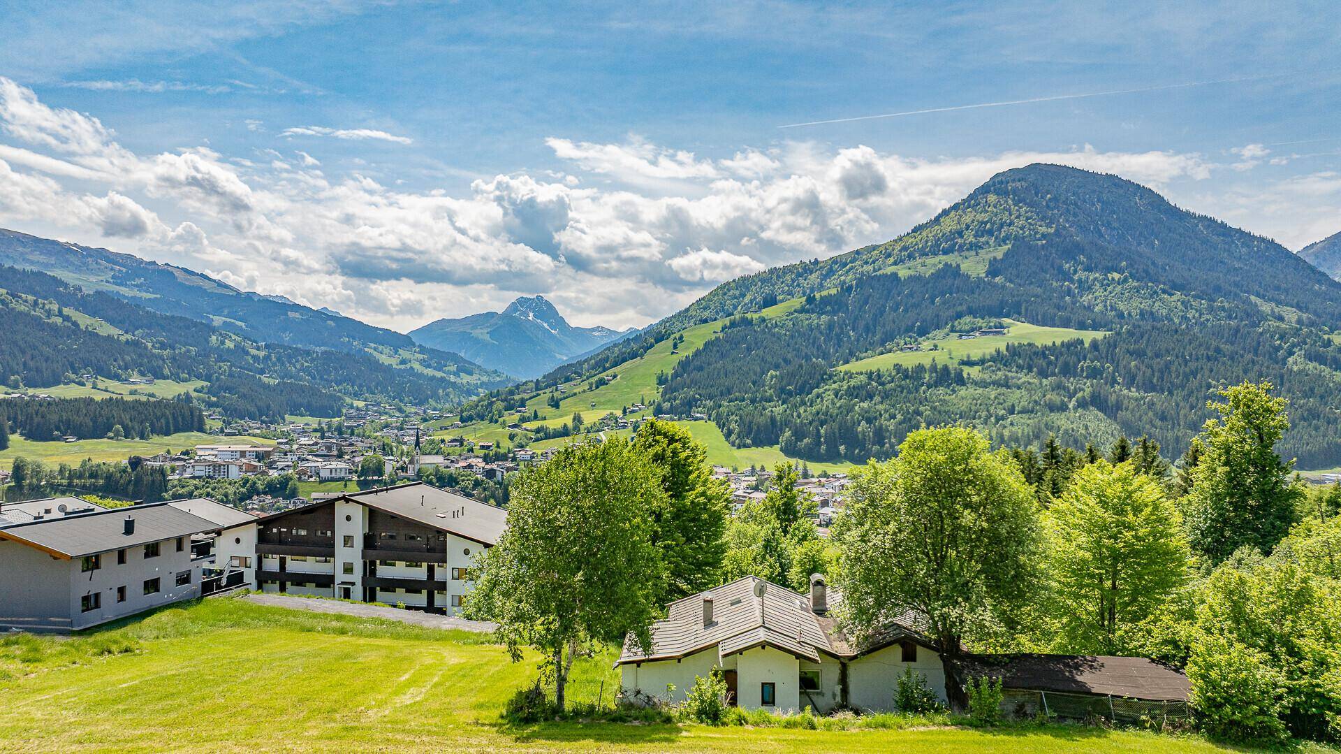 Weitläufiger Blick auf eine Siedlung in den Bergen, umgeben von grünen Wiesen und Wäldern.