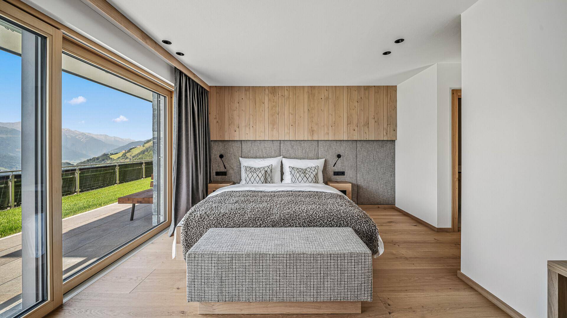 Helles Schlafzimmer mit Holzwand, großem Bett und Zugang zur Terrasse mit Bergblick.