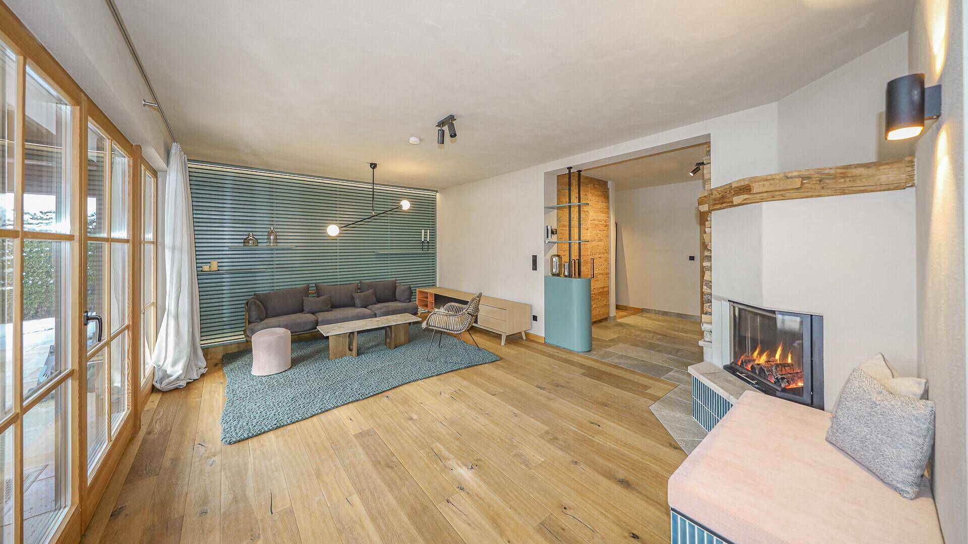 Gemütliches Wohnzimmer mit Holzboden, Kamin und großen Fenstern, die viel Licht hereinlassen.