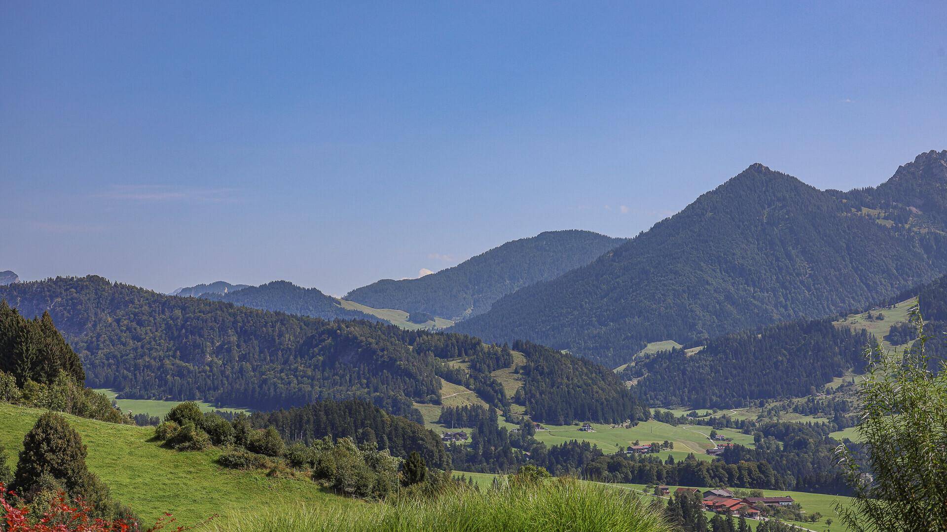 Weitläufige Landschaft mit sanften grünen Hügeln, Tälern und vereinzelten Häusern in der Ferne.