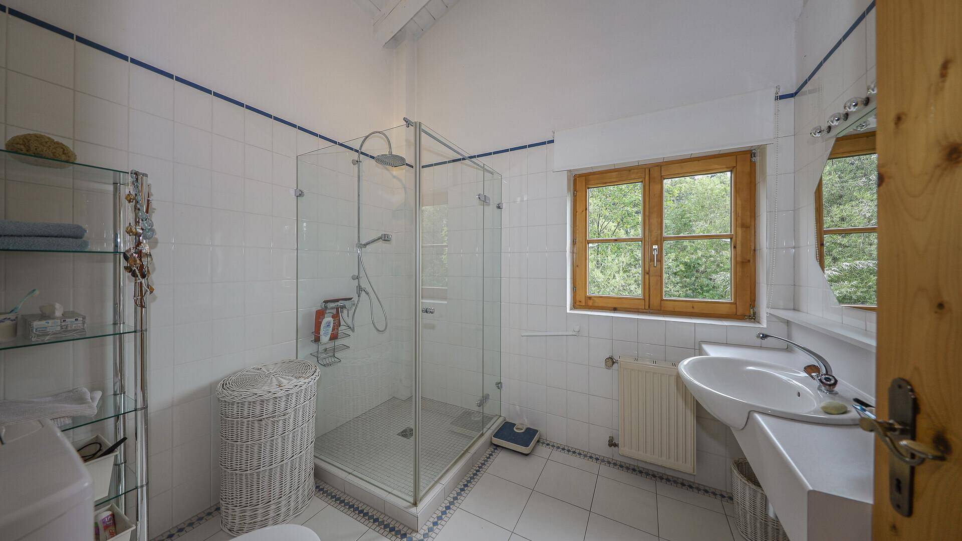 Helles Badezimmer mit weißen Fliesen, Duschkabine und Fenster mit Blick ins Grüne.