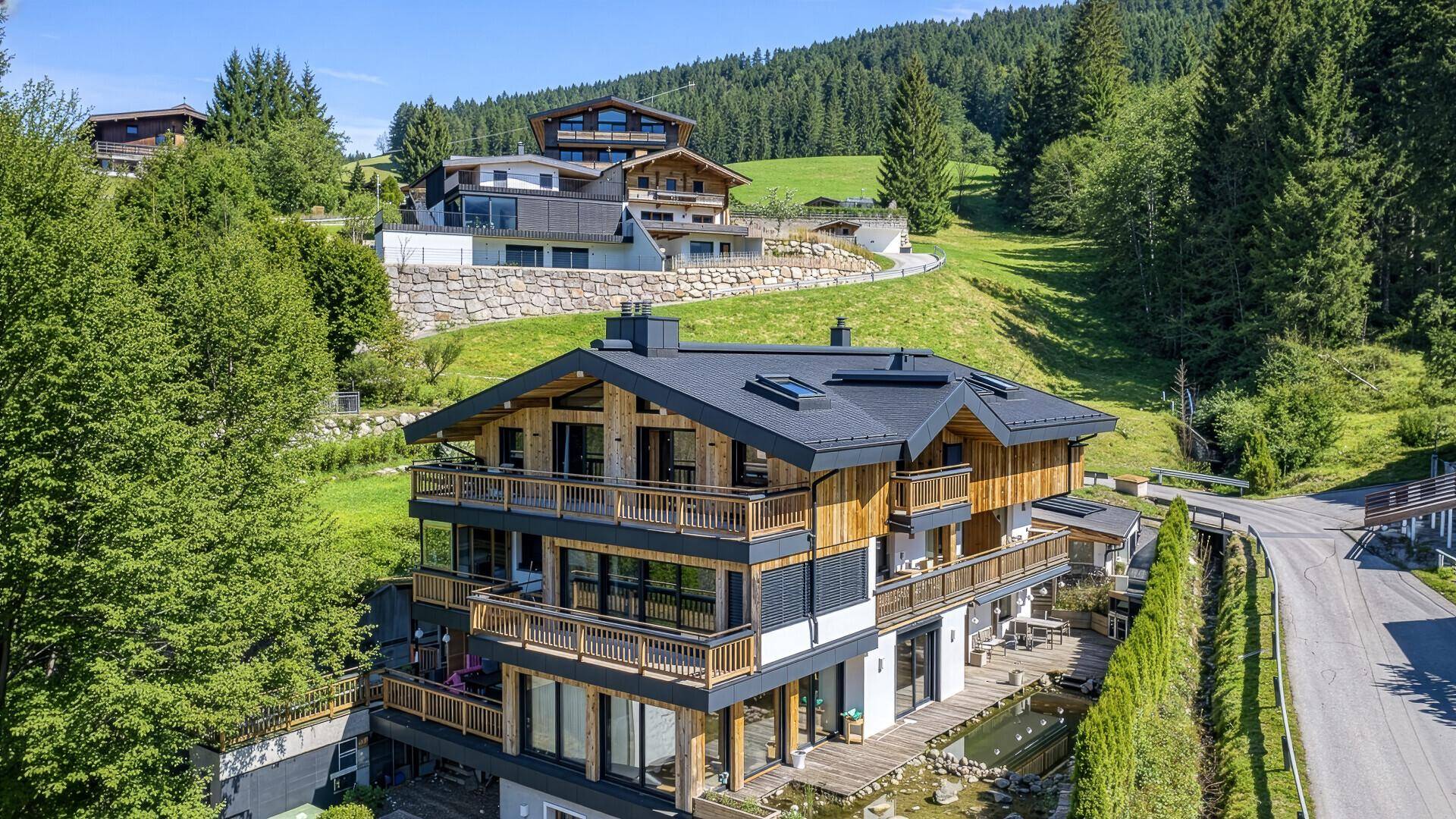 Luxuriöses Chalet in alpiner Umgebung mit Wald und weiteren Gebäuden unter blauem Himmel.